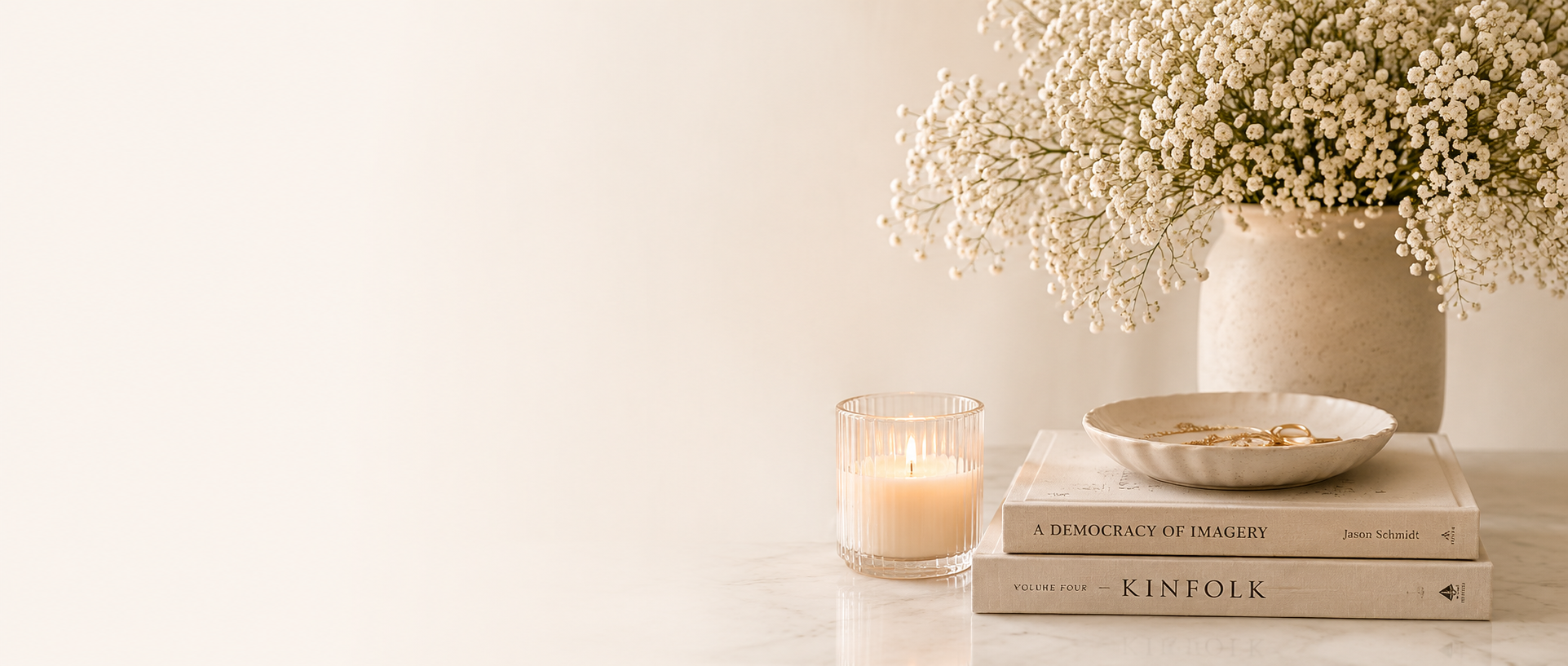 A lit candle beside a stack of books topped with a small dish of jewelry on a marble surface