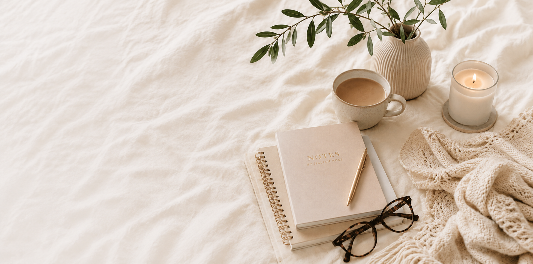A notebook, mug of coffee, lit candle, and vase of greenery styled on a linen background